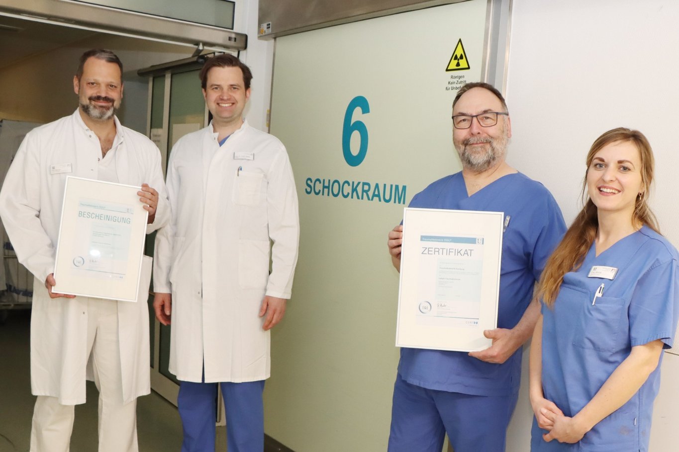 Vier medizinische Fachkräfte stehen vor dem Schockraum eines Krankenhauses und halten Zertifikate in den Händen.