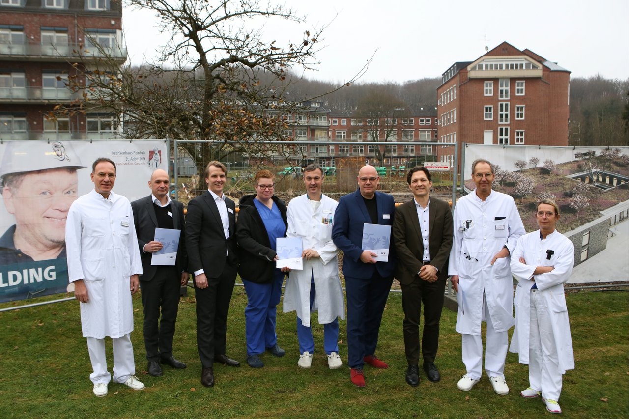 Gruppe von mehreren Personen, darunter medizinisches Personal und Personen in Anzügen, stehen im Freien vor dem Krankenhaus und halten Dokumente in den Händen.