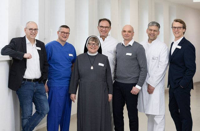 Sieben Personen stehen in einem Flur nebeneinander und lächeln. 