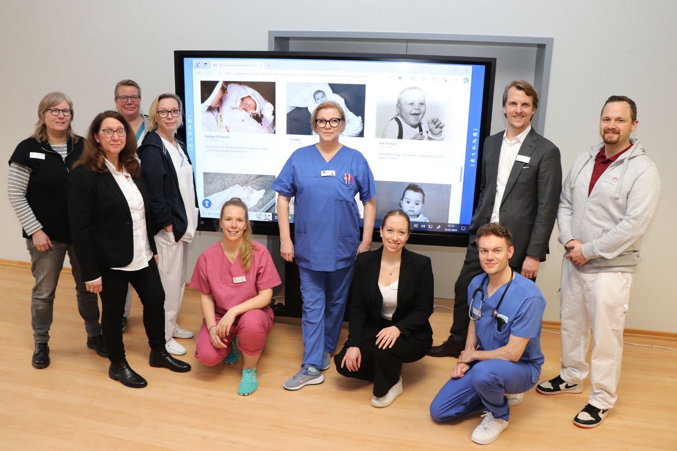 Eine Gruppe von Mitarbeitenden des St. Adolf-Stifts aus verschiedenen Berufsbereichen steht vor einem Smartboard mit Babyfotos aus unterschiedlichen Jahrzehnten.