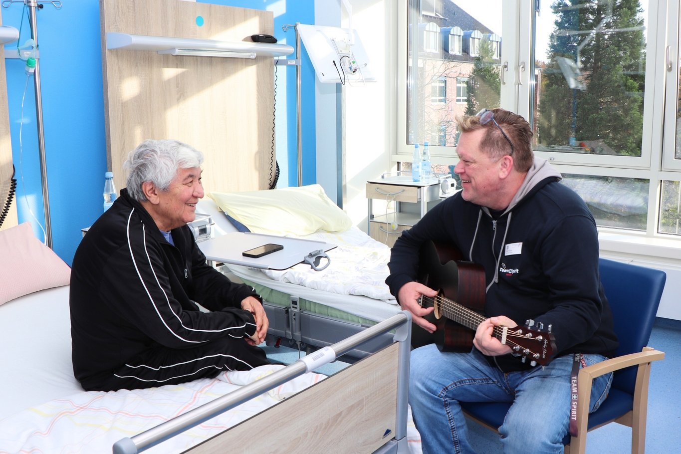 Ein Patient sitzt auf seinem Krankenhausbett und hört einem Mann zu, der ihm in einem hellen Patientenzimmer auf einer Gitarre vorspielt. Beide lächeln sich an.