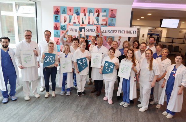 Großes Team aus medizinischem Personal im Krankenhaus Reinbek steht gemeinsam in einem Flur und hält Zertifikate sowie Schilder hoch, im Hintergrund ein Banner mit der Aufschrift „Danke – wir sind ausgezeichnet!“.