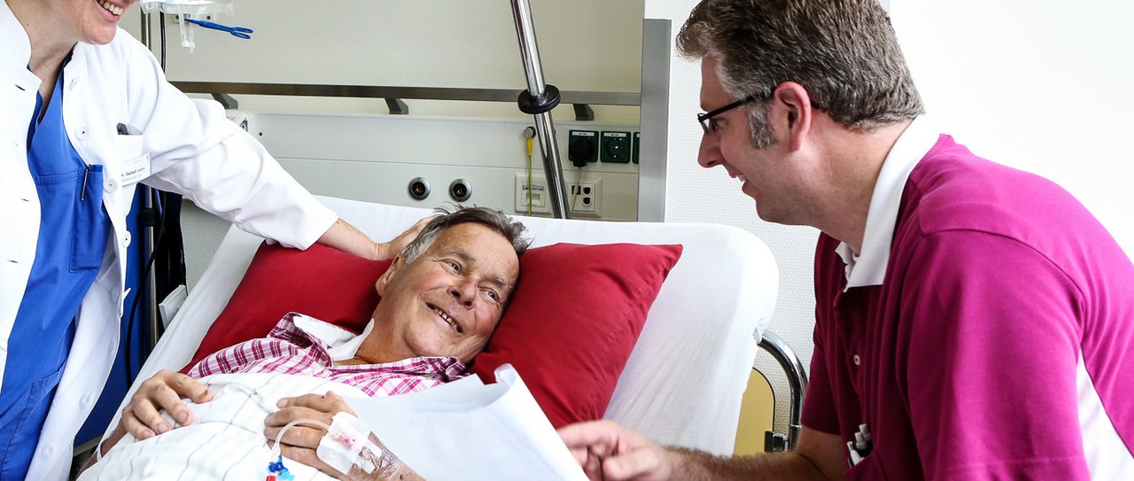 Ein Dialyse-Patient liegt im Krankenhausbett und ein Arzt sitzt daneben und redet mit ihm