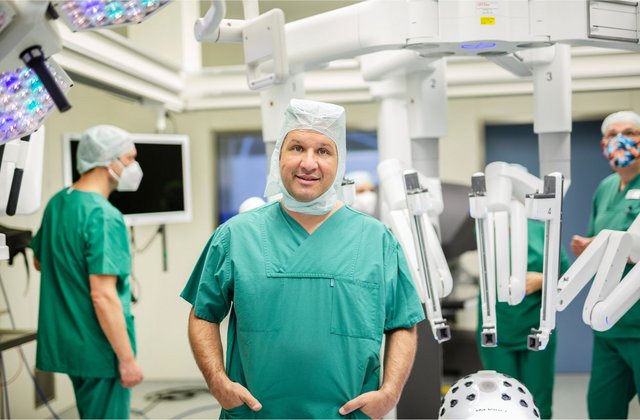 Arzt in grüner OP-Kleidung steht in einem Operationssaal neben einem da-Vinci-Operationsroboter, während ein OP-Team im Hintergrund vorbereitet ist.