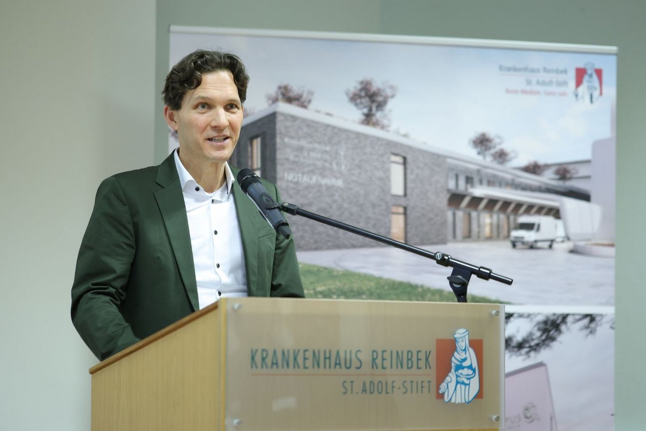 Ein Mann im Anzug steht am Rednerpult mit Mikrofon und hält einen Vortrag vor einer Präsentationswand im Krankenhaus Reinbek.