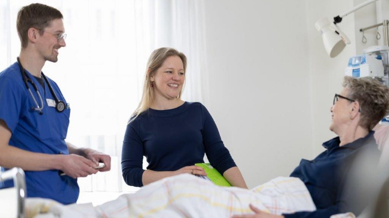 Ein Pfleger und eine Mitarbeiterin des Sozialdientes sprechen mit einer Neurologiepatientin am Krankenhausbett