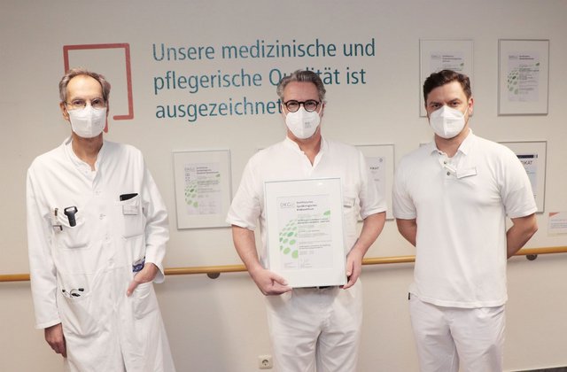 Drei Ärzte mit Mundschutz stehen vor einer Wand mit Zertifikaten, während einer von ihnen ein Zertifikat für das gynäkologische Krebszentrum in den Händen hält.