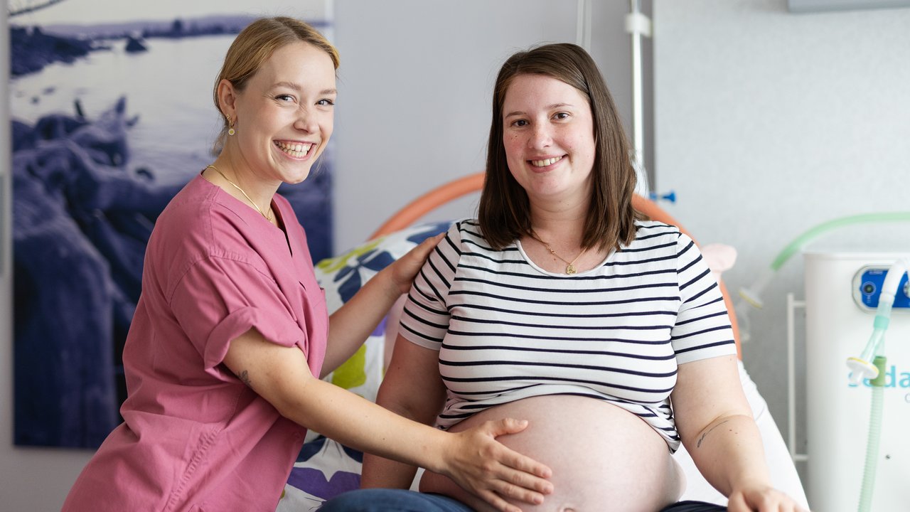 Eine Hebamme hält den Bauch einer Schwangeren. Beide lächeln in die Kamera