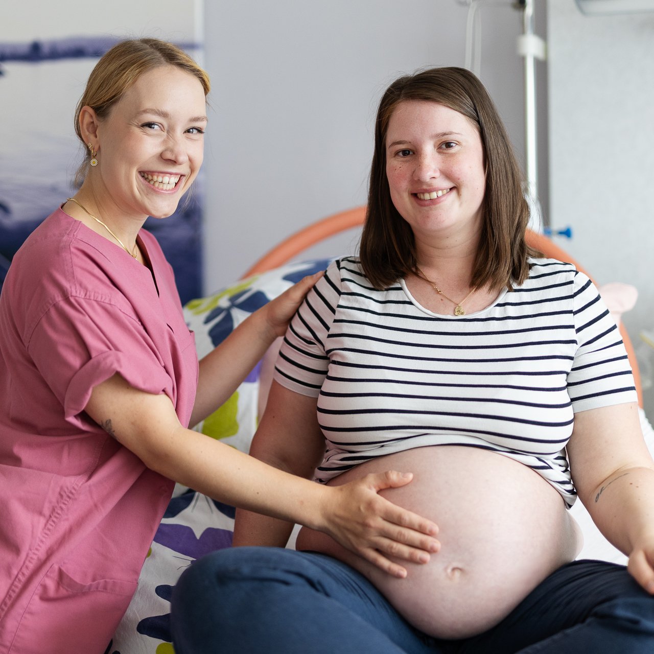 Eine Hebamme hält den Bauch einer Schwangeren. Beide lächeln in die Kamera