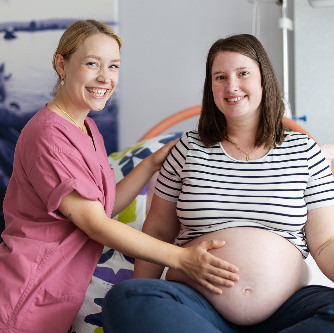 Eine Hebamme hält den Bauch einer Schwangeren. Beide lächeln in die Kamera