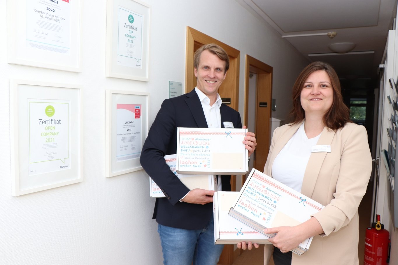 Zwei Personen, ein Mann und eine Frau in Businesskleidung, stehen in einem Flur und halten mehrere Geschenkboxen mit der Aufschrift „Bester Start ins Leben“ in den Händen.