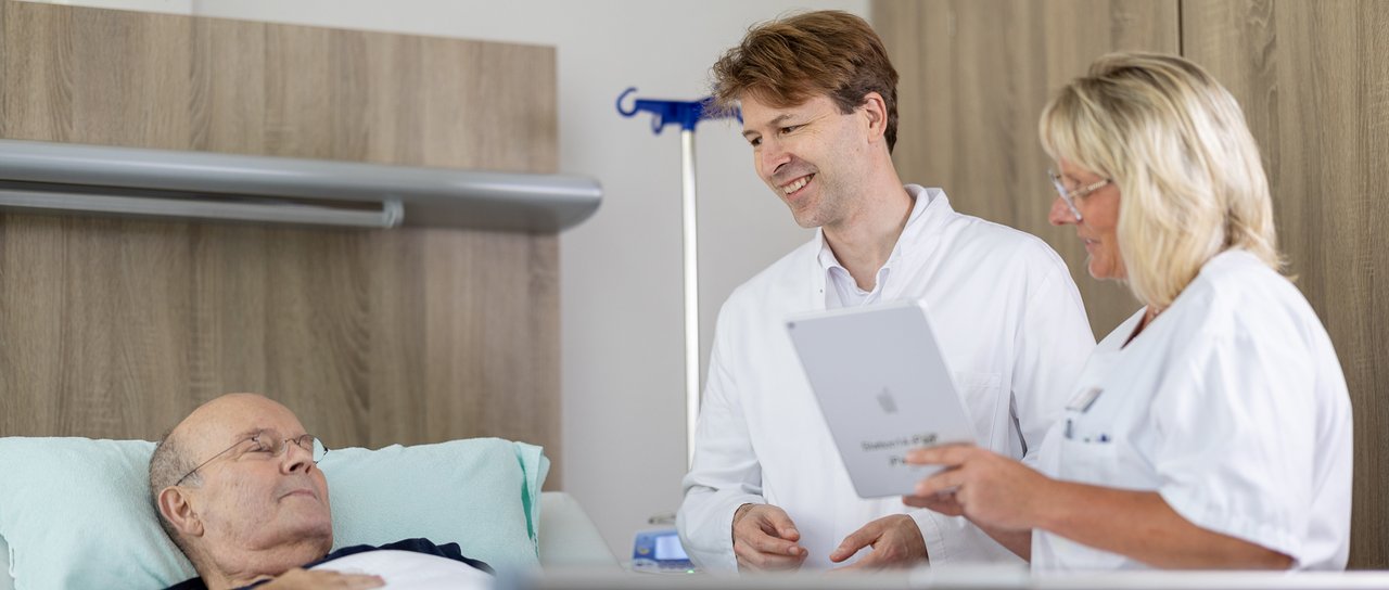 Ein Arzt und eine Pflegerin stehen am Patentenbett eines älteren Herren und sprechen mit ihm.