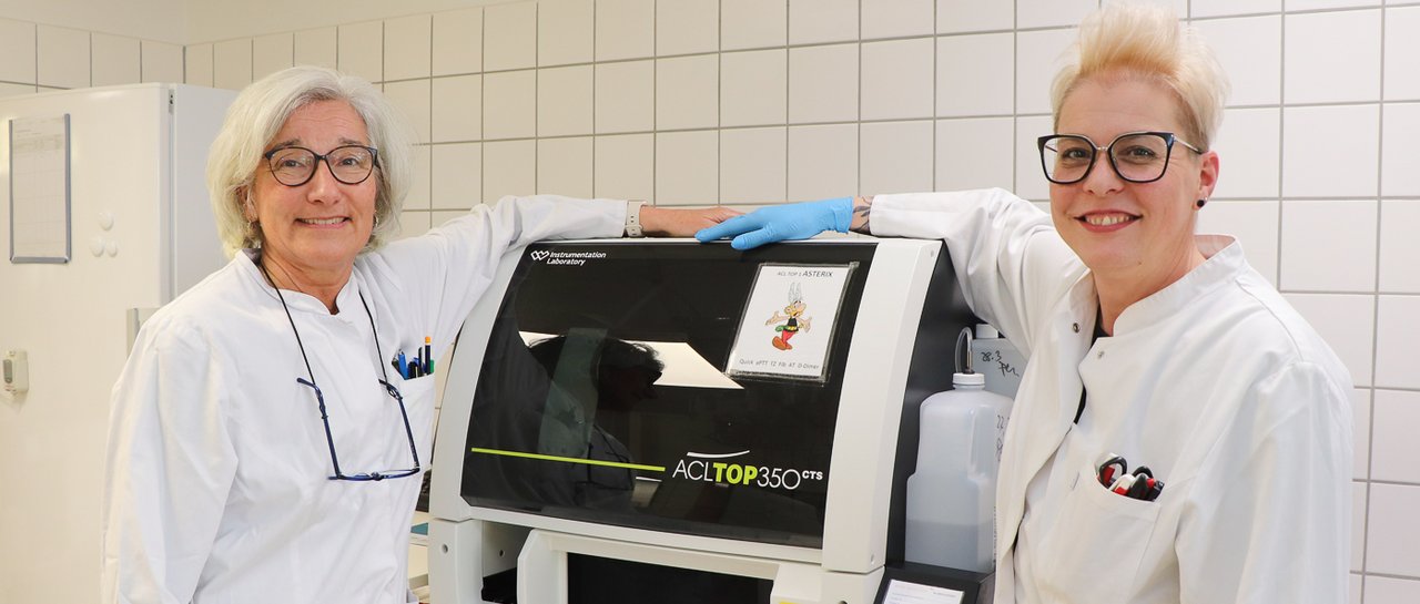 Zwei Laborantinnen stehen vor einem Gerät und lächeln in die Kamera