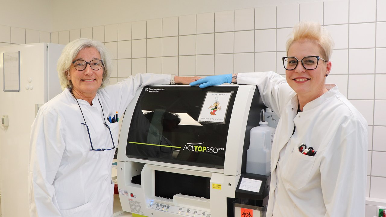Zwei Laborantinnen stehen vor einem Gerät und lächeln in die Kamera