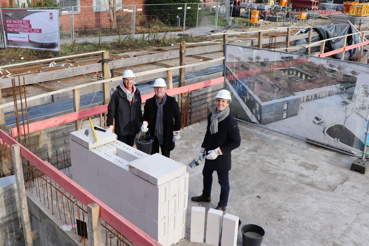 Drei Personen mit Bauhelmen stehen auf einer Baustelle neben einer neu gemauerten Wand und halten Werkzeug in den Händen.