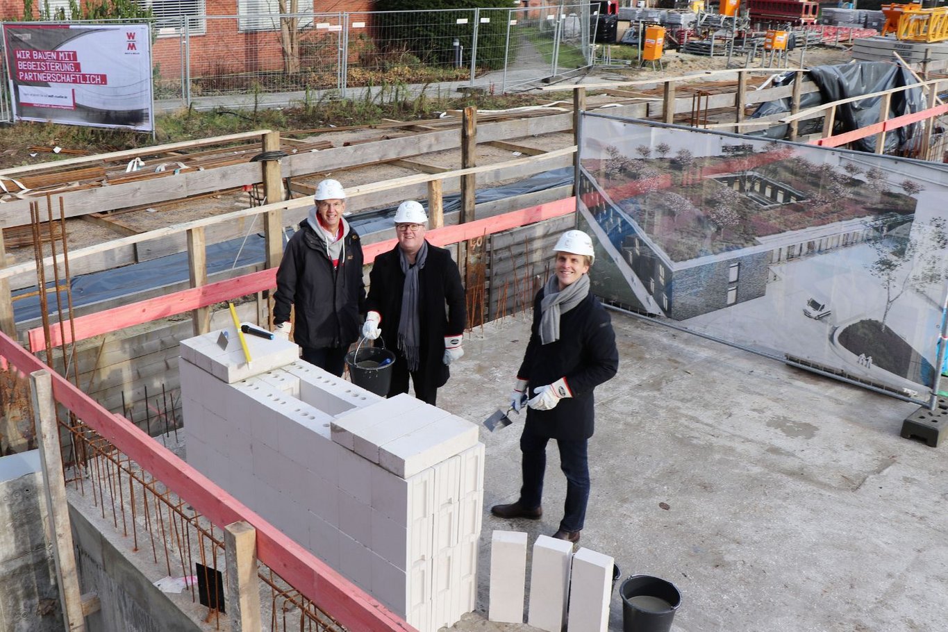 Drei Personen mit Bauhelmen mauern auf einer Baustelle eine Wand und beteiligen sich an der symbolischen Grundsteinlegung eines Neubaus am Krankenhaus Reinbek.