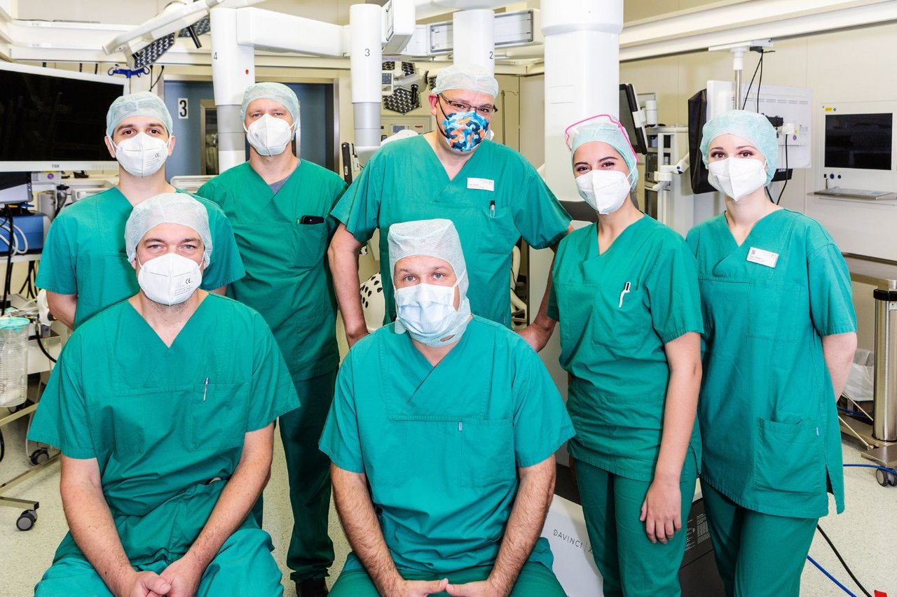 Mehrere Mitglieder eines OP-Teams in grüner Schutzkleidung stehen gemeinsam im Operationssaal vor medizinischen Geräten.