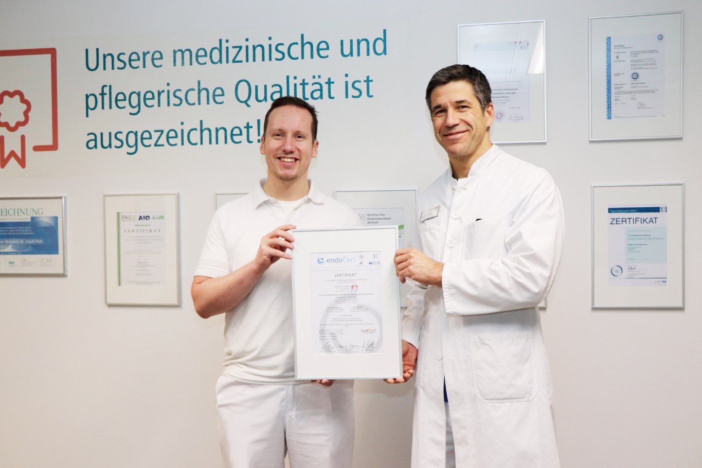 Zwei Ärzte in weißer Arbeitskleidung stehen nebeneinander in einem Klinikflur und halten gemeinsam ein gerahmtes Zertifikat. Im Hintergrund sind mehrere an der Wand befestigte Urkunden und der Schriftzug unsere medizinische und pflegerische Qualität ist ausgezeichnet zu sehen.