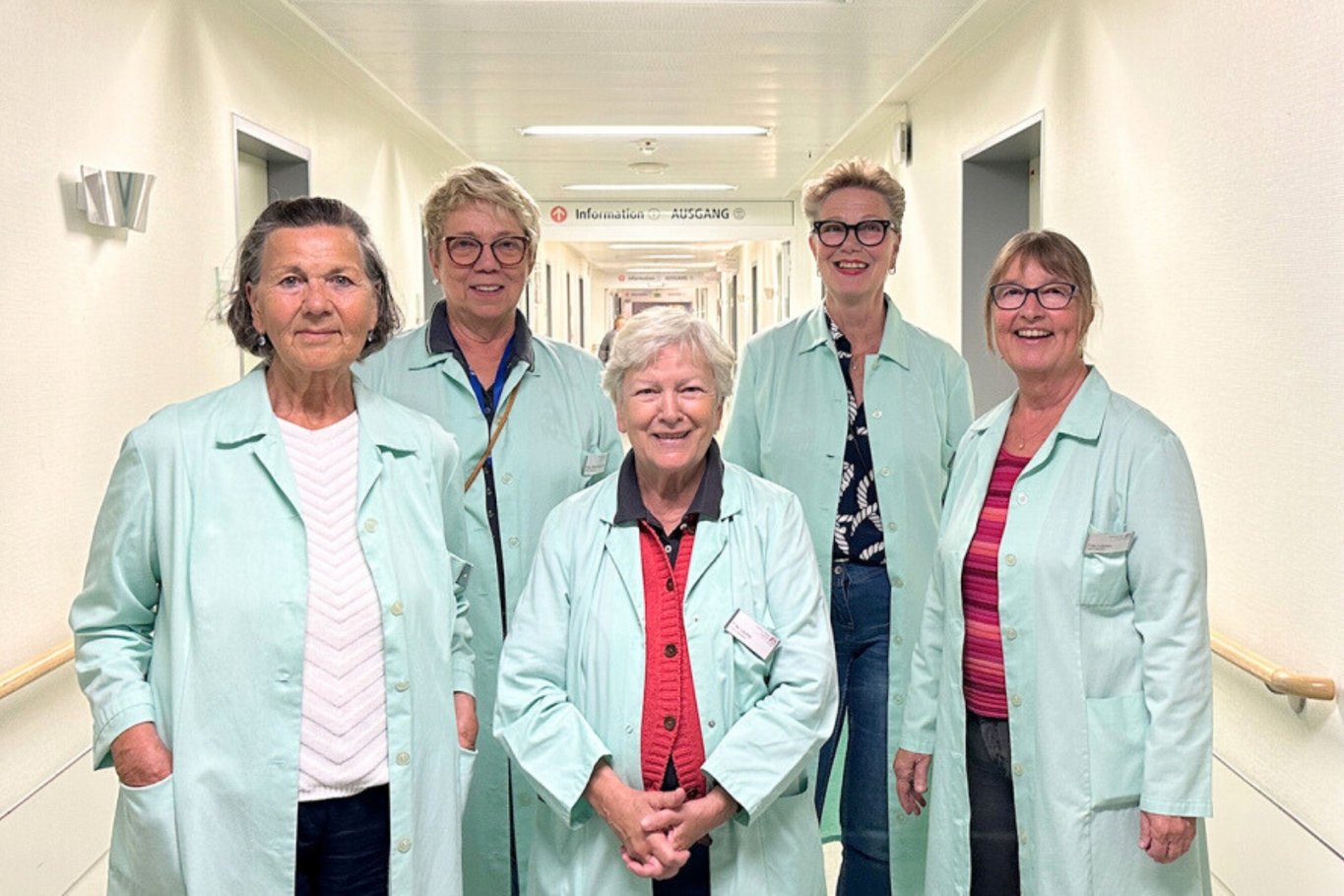 Fünf Frauen in hellgrünen Kitteln stehen gemeinsam auf einem Flur im Krankenhaus. Sie blicken mit einem Lächeln in die Kamera.