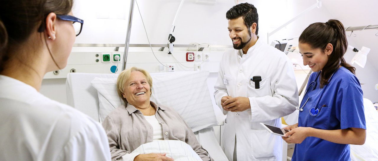 Ärzte und Pflegekräfte stehen um das Bett einer älteren Patientin. Alle unterhalten sich angeregt