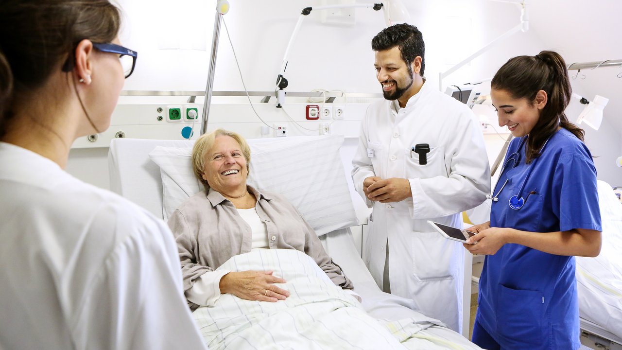 Ärzte und Pflegekräfte stehen um das Bett einer älteren Patientin. Alle unterhalten sich angeregt