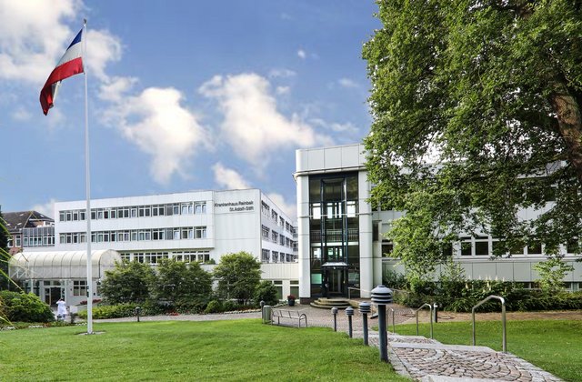 Außenansicht des Krankenhauses Reinbek St. Adolf-Stift mit Fahnenmast, Grünfläche und Eingangsbereich bei sonnigem Wetter.