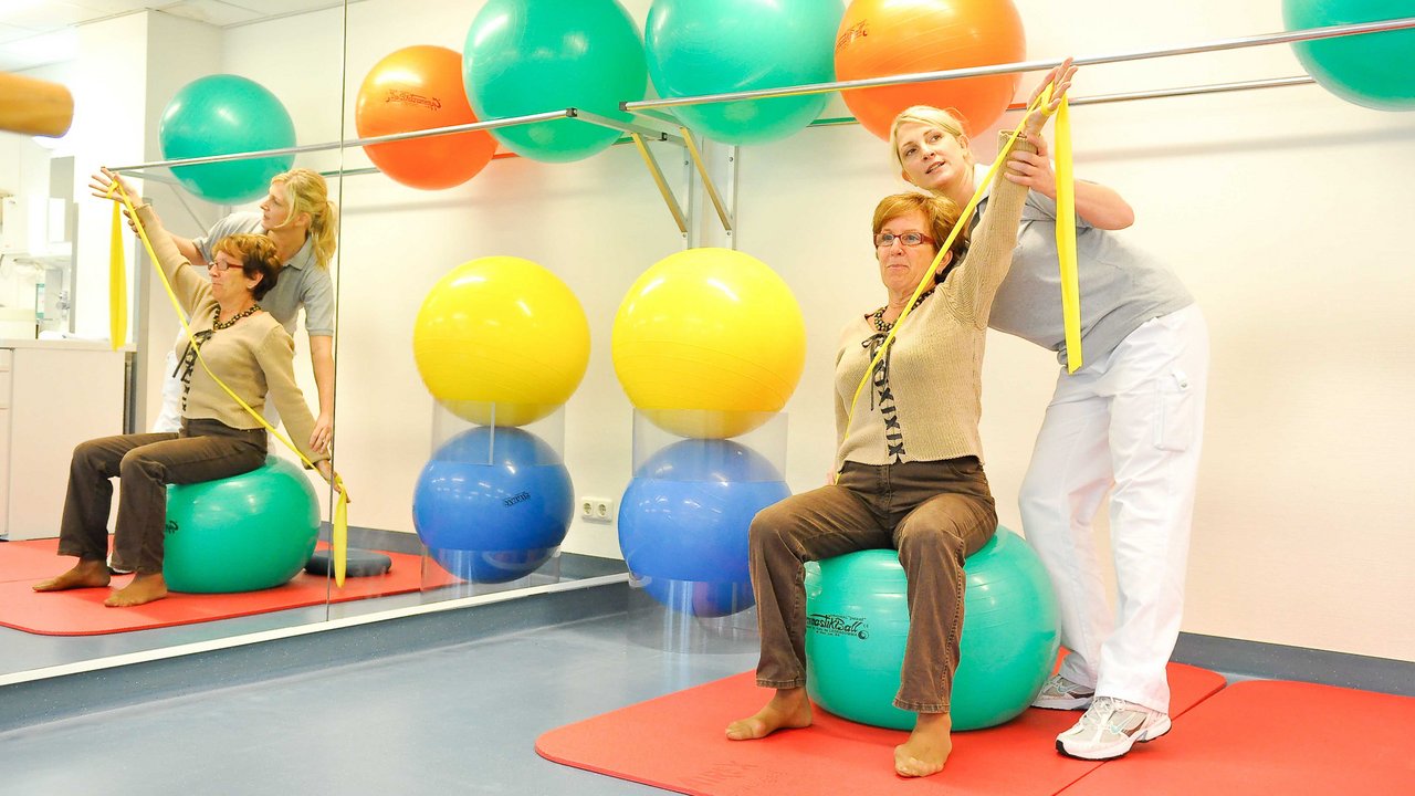 Eine Dame macht mit einer Therapeutin eine Übung auf einem Gymastikball