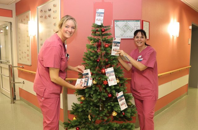 Zwei Pflegekräfte in pinker Arbeitskleidung schmücken einen Weihnachtsbaum auf einer Station im Krankenhaus Reinbek mit Broschüren und Weihnachtsdekoration.