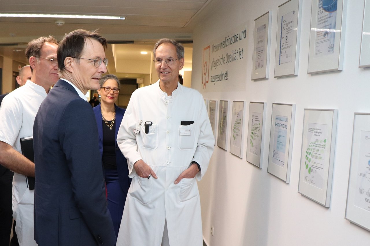 Mehrere Personen, darunter ein Mann im Anzug und Ärzte in weißer Kleidung, betrachten gemeinsam Zertifikate an einer Wand im Krankenhaus Reinbek.