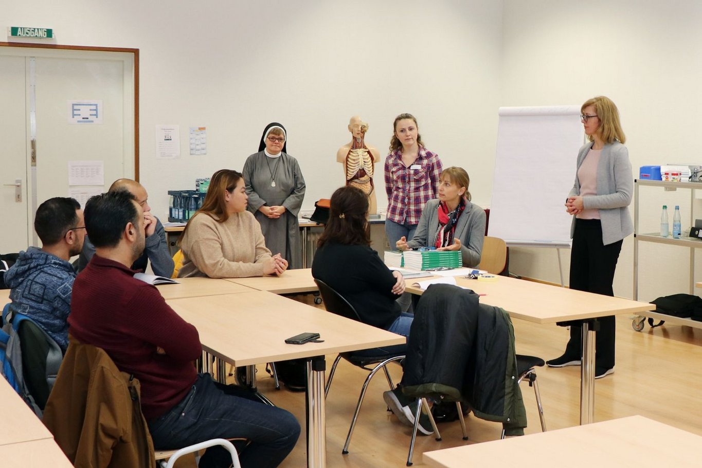 Mehrere Kursteilnehmende sitzen in einem Klassenraum an Tischen und hören den Dozentinnen zu, die vorne stehen und den Unterricht eines berufsbezogenen Sprachkurses leiten; im Hintergrund sind ein Flipchart und ein Anatomiemodell zu sehen.
