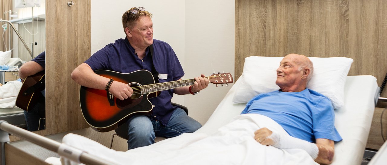 Ein man sitzt am Krankenhausbett eines Patienten und spielt ihm ein Lied auf der Gitarre vor