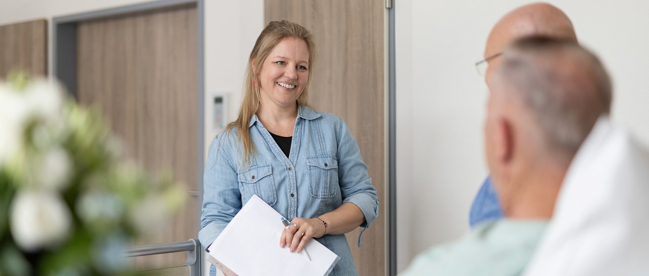 Eine Sozialdienst-Mitarbeiterin hält Zettel in den Händen und berät ein Paar am Krankenhausbett