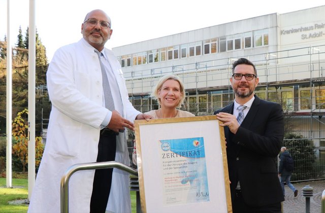 Drei Personen stehen vor dem Gebäude des Krankenhauses Reinbek St. Adolf-Stift und halten ein Zertifikat der DGAV für das Kompetenzzentrum für Magen- und Speiseröhrenchirurgie.