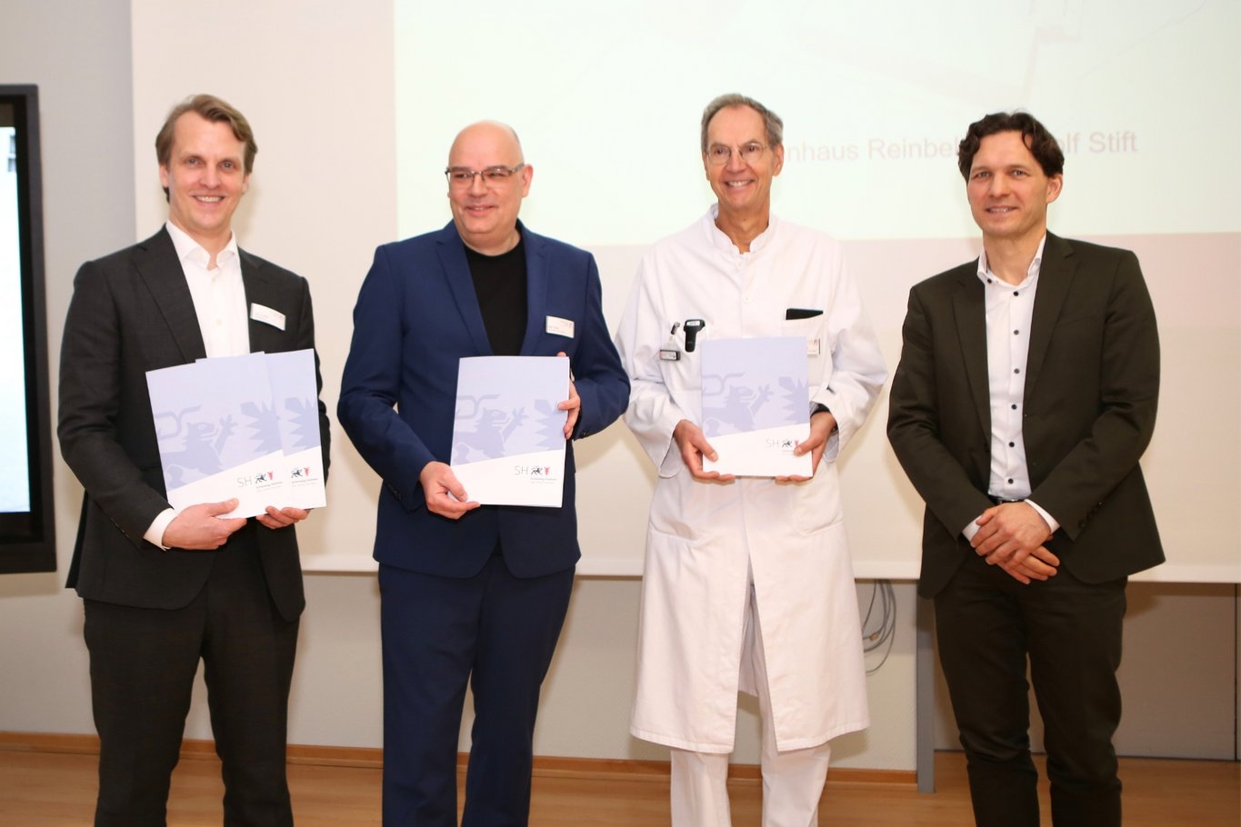Vier Männer stehen nebeneinander und halten Förderbescheide in den Händen, die im Rahmen einer offiziellen Übergabe für Bauprojekte des Krankenhauses Reinbek überreicht wurden.