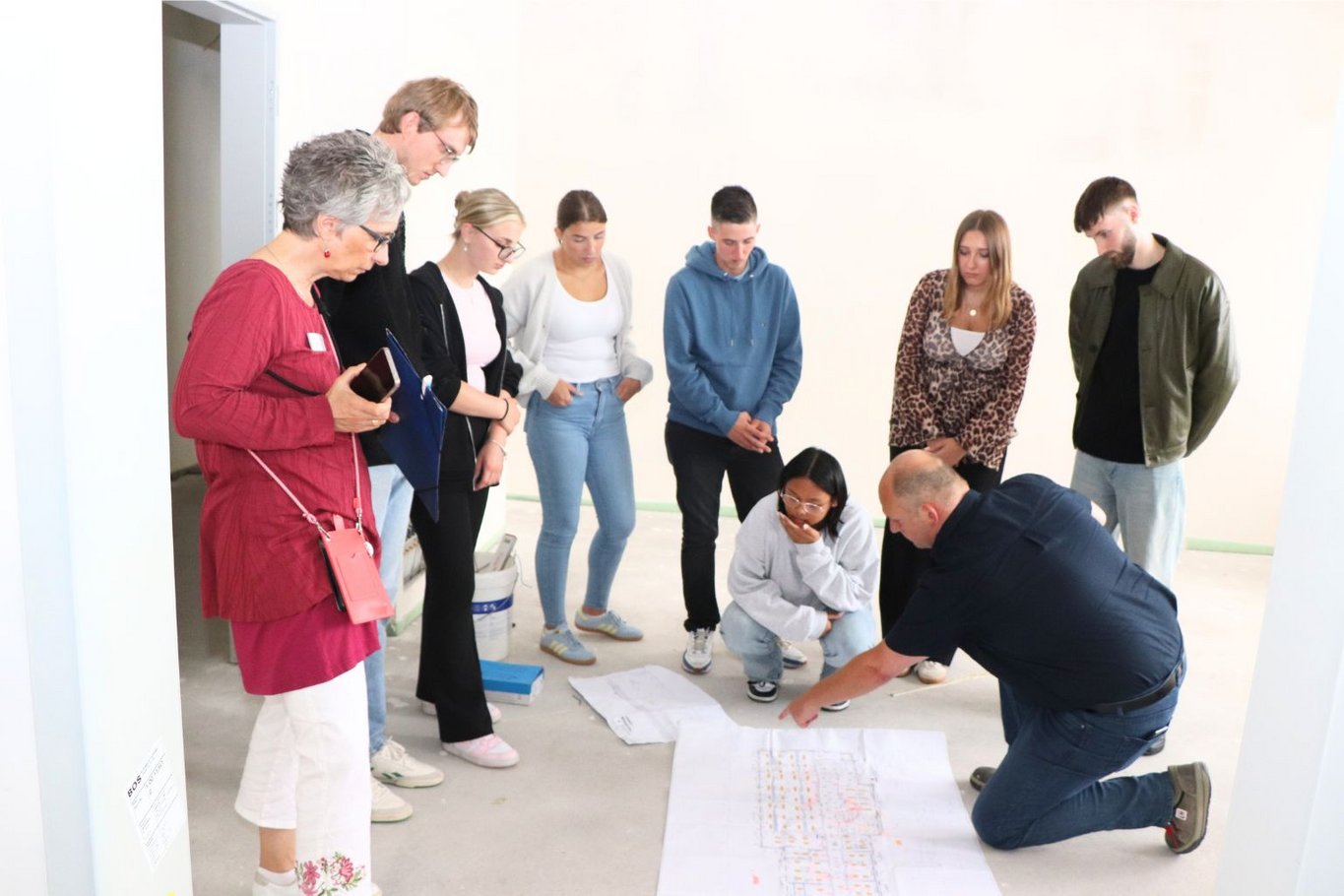 Gruppe von Auszubildenden und zwei Leitungen stehen in einem Raum des neuen Campus um ausgebreitete Baupläne, während ein Mann darauf zeigt und etwas erklärt.