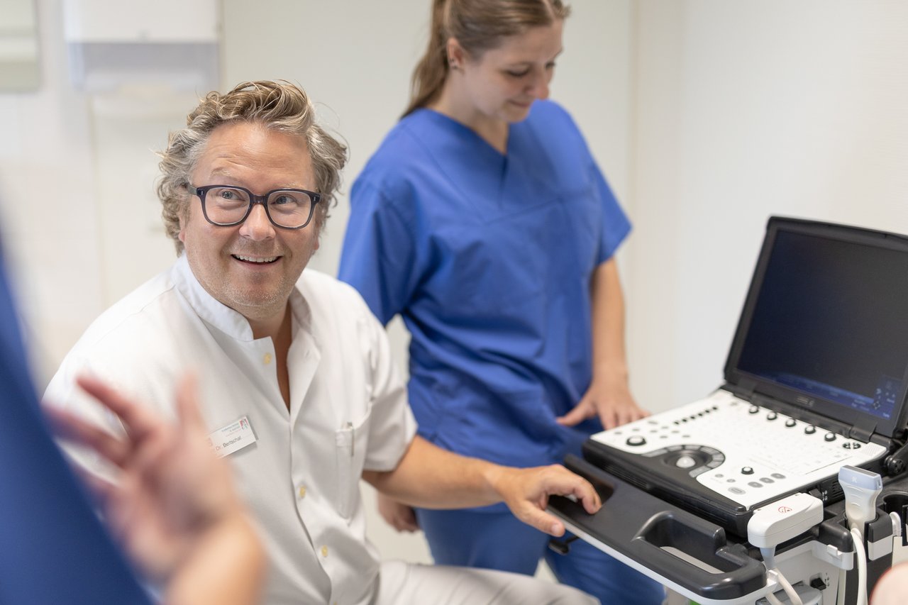 Ein Arzt sitzt an einem Ultraschallbildschirm und lächelt, im Hintergrund eine Pflegekraft