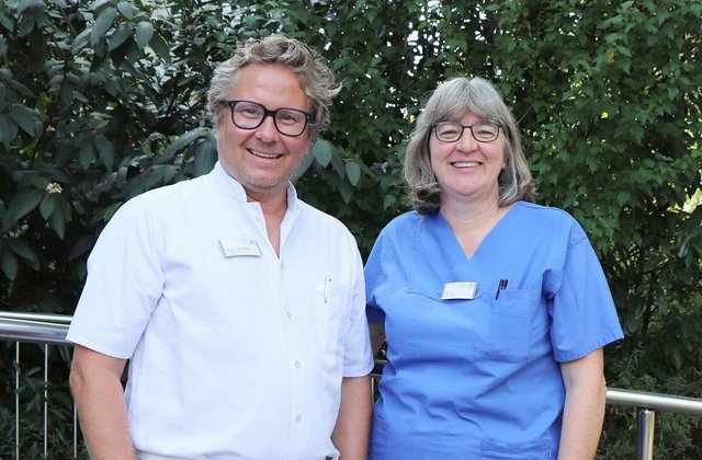 Zwei Ärzt:innen des St. Adolf-Stiftes stehen nebeneinander im Freien vor grüner Vegetation. Links ein Mann in weißem Arztkittel, rechts eine Frau in blauer medizinischer Arbeitskleidung. Beide tragen Namensschilder und lächeln in die Kamera.