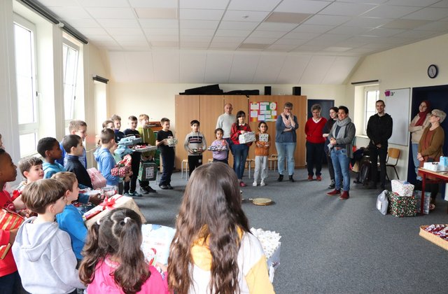 Kinder stehen in einem Klassenraum im Kreis, halten Weihnachtsgeschenke in den Händen und freuen sich gemeinsam, während mehrere Erwachsene am Rand zuschauen.