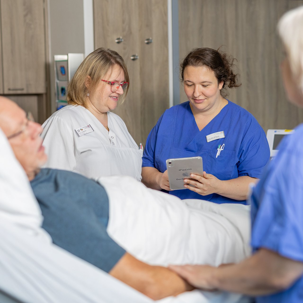 Ein älterer Patient liegt im Patientenbett eine Pflegerin und eine Ärztin tauschen sich aus und schauen auf ein Tablet