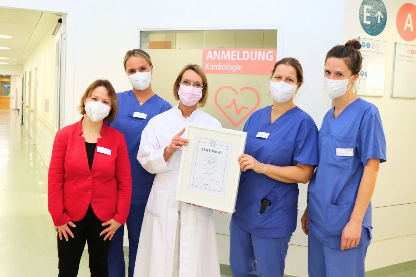 Fünf Mitarbeiterinnen eines Krankenhauses stehen vor der Anmeldung der Kardiologie und präsentieren gemeinsam ein Zertifikat für die Chest Pain Unit.