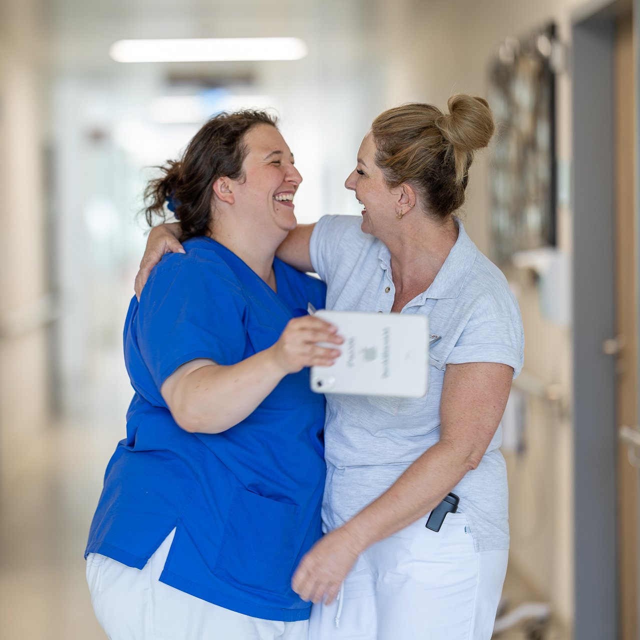 Zwei Pflegekräfte lachen und umarmen sich herzlich auf dem Krankenhausflur