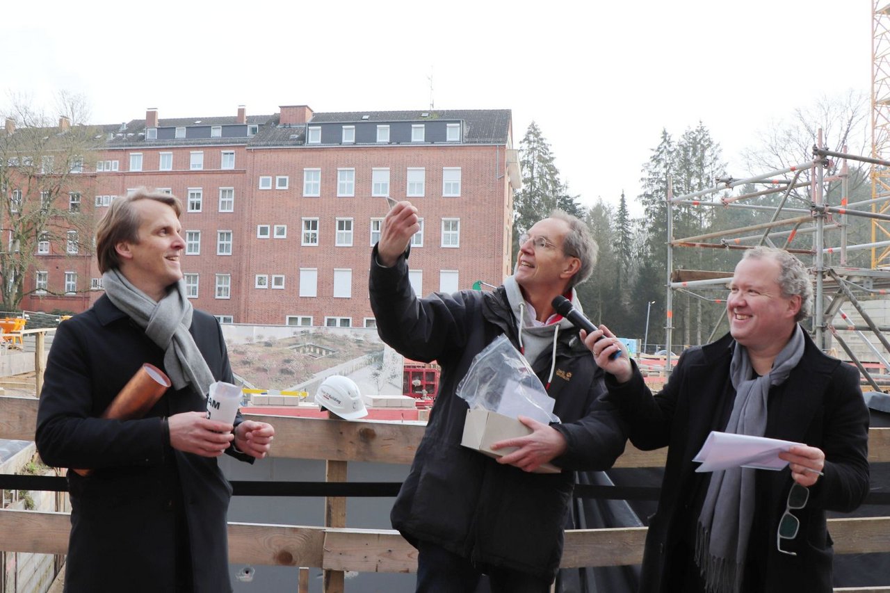 Drei Männer stehen auf der Baustelle, einer hält ein Mikrofon und spricht, während die anderen zuhören.