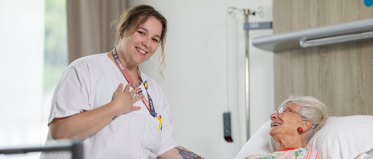 Eine Pflegerin hält die Hand einer Patientin, welche im Krankenhausbett liegt. Die Pflegerin hält ihre andere Hand auf ihr Herz und lacht in die Kamera