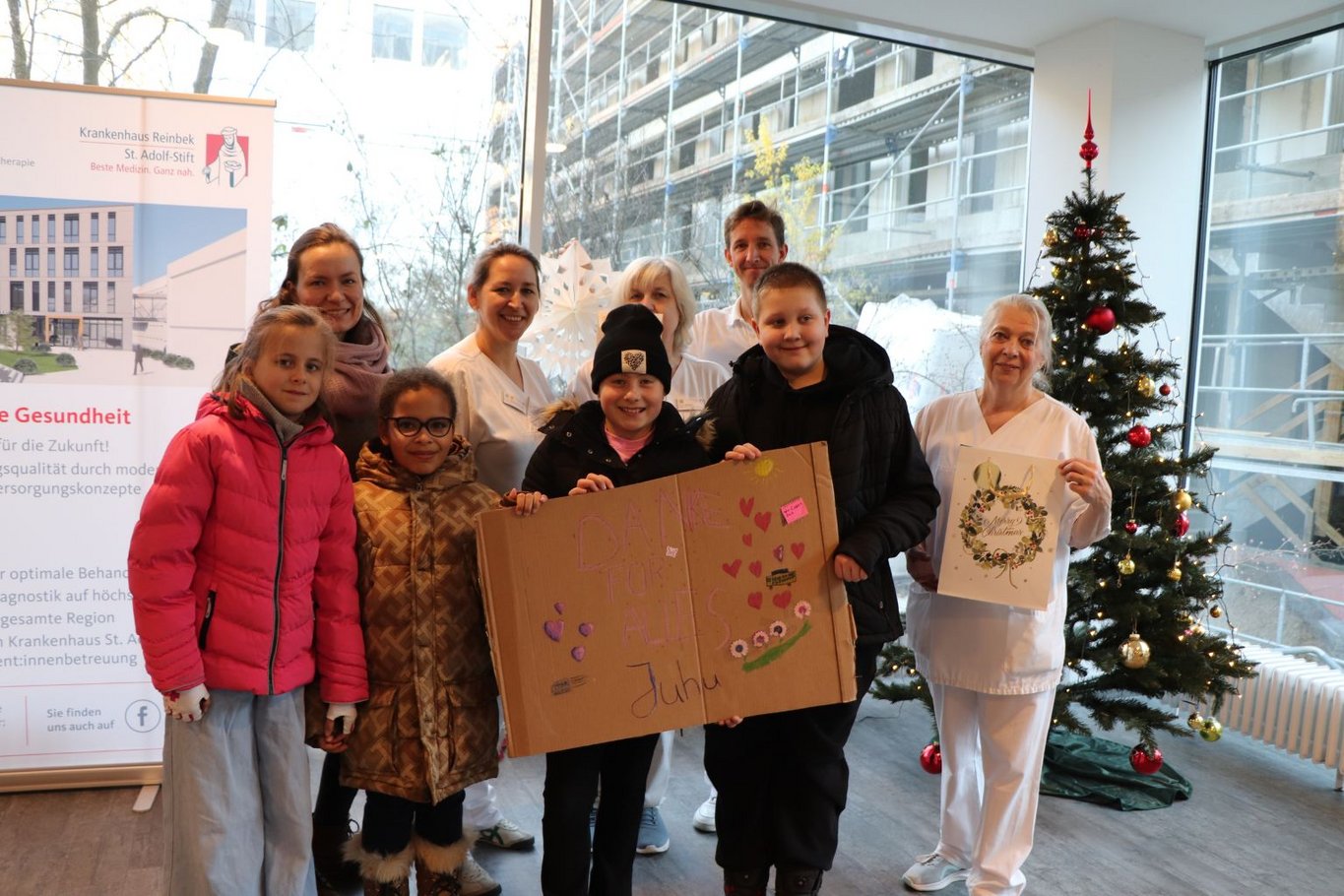 Mehrere Kinder und Erwachsene stehen gemeinsam in einem Krankenhaus vor einem Weihnachtsbaum; die Kinder halten ein selbst gestaltetes Plakat und Geschenke für die Mitarbeitenden.