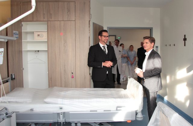 Zwei Männer stehen in einem neu eingerichteten Patientenzimmer mit Klinikbett und moderner Ausstattung und sprechen miteinander, während im Hintergrund Klinikpersonal im Flur steht.