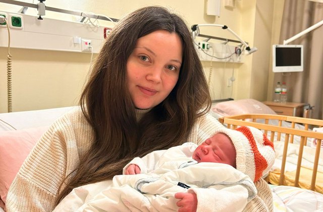 Mutter sitzt in einem Krankenhauszimmer und hält ihr neugeborenes Baby im Arm, das warm eingepackt ist und eine Mütze trägt.