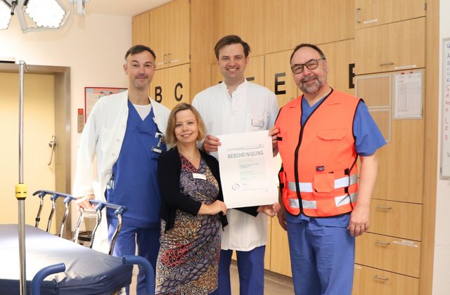 Vier Mitarbeitende des Krankenhauses stehen im Schockraum neben einer Behandlungsliege und halten ein Zertifikat zur erneuten Auszeichnung als lokales Traumazentrum in die Kamera.