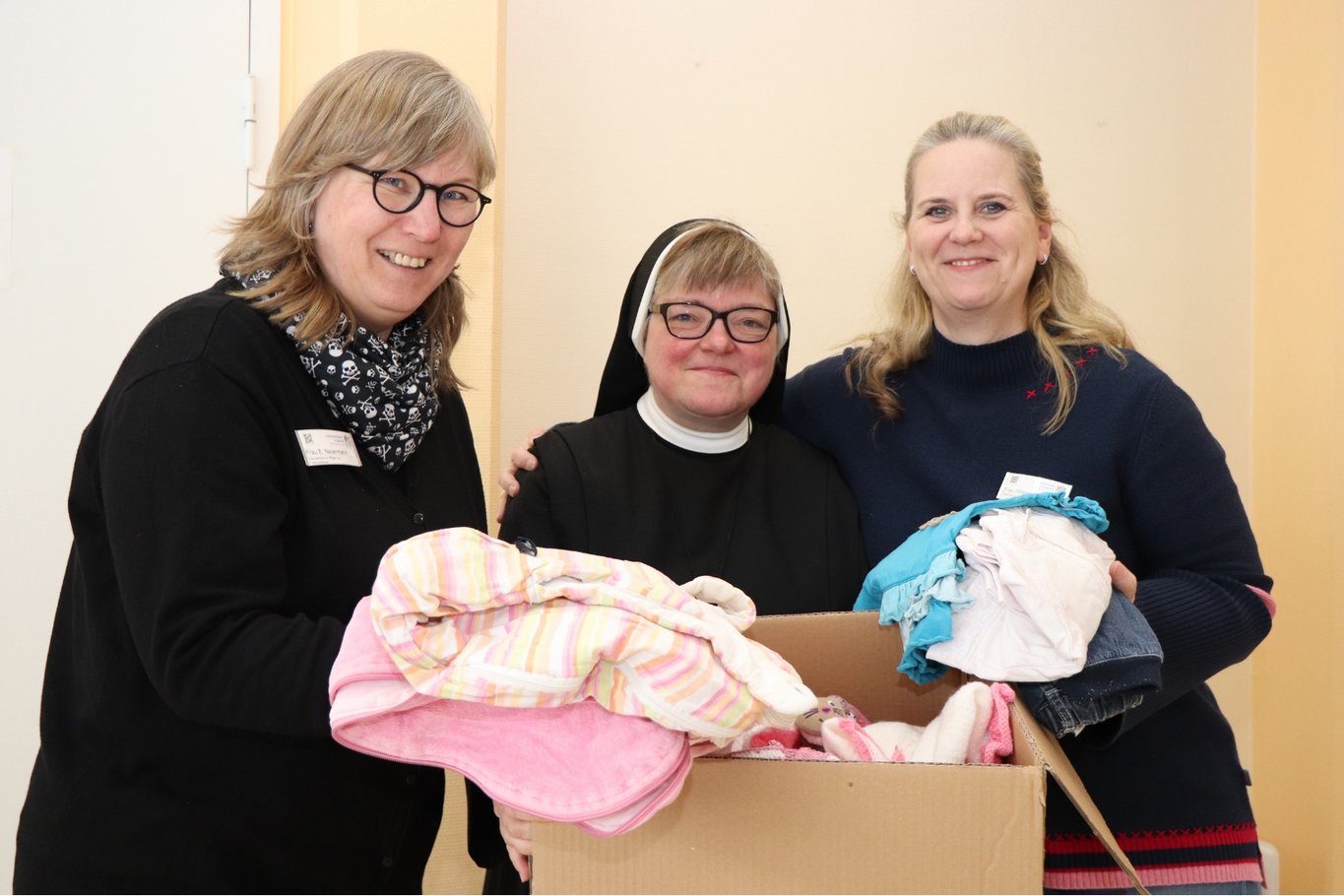 Drei Frauen stehen nebeneinander und halten eine Kiste mit Hilfsgütern in der Hand.