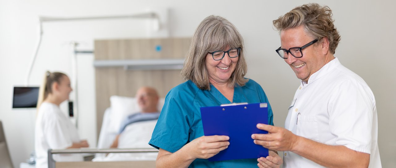 Dr. Dick und Bertschart stehen vor einem Patientenbett und besprechen etwas, das sie auf dem Tablet sehen