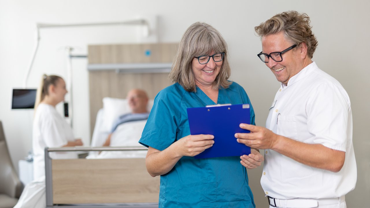 Dr. Dick und Bertschart stehen vor einem Patientenbett und besprechen etwas, das sie auf dem Tablet sehen